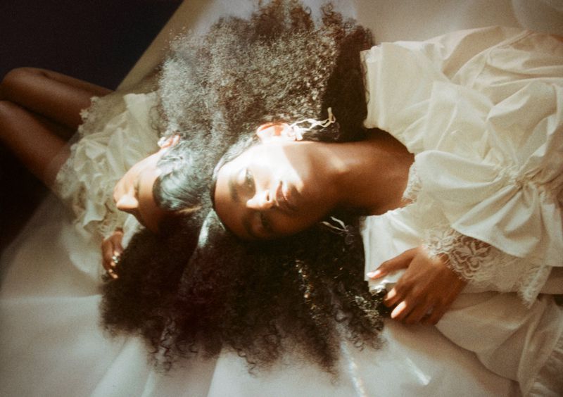 Model Tsion Teferi lying head-to-head wearing an ivory ruffled lace dress with voluminous dark curly hair spreading across white fabric.