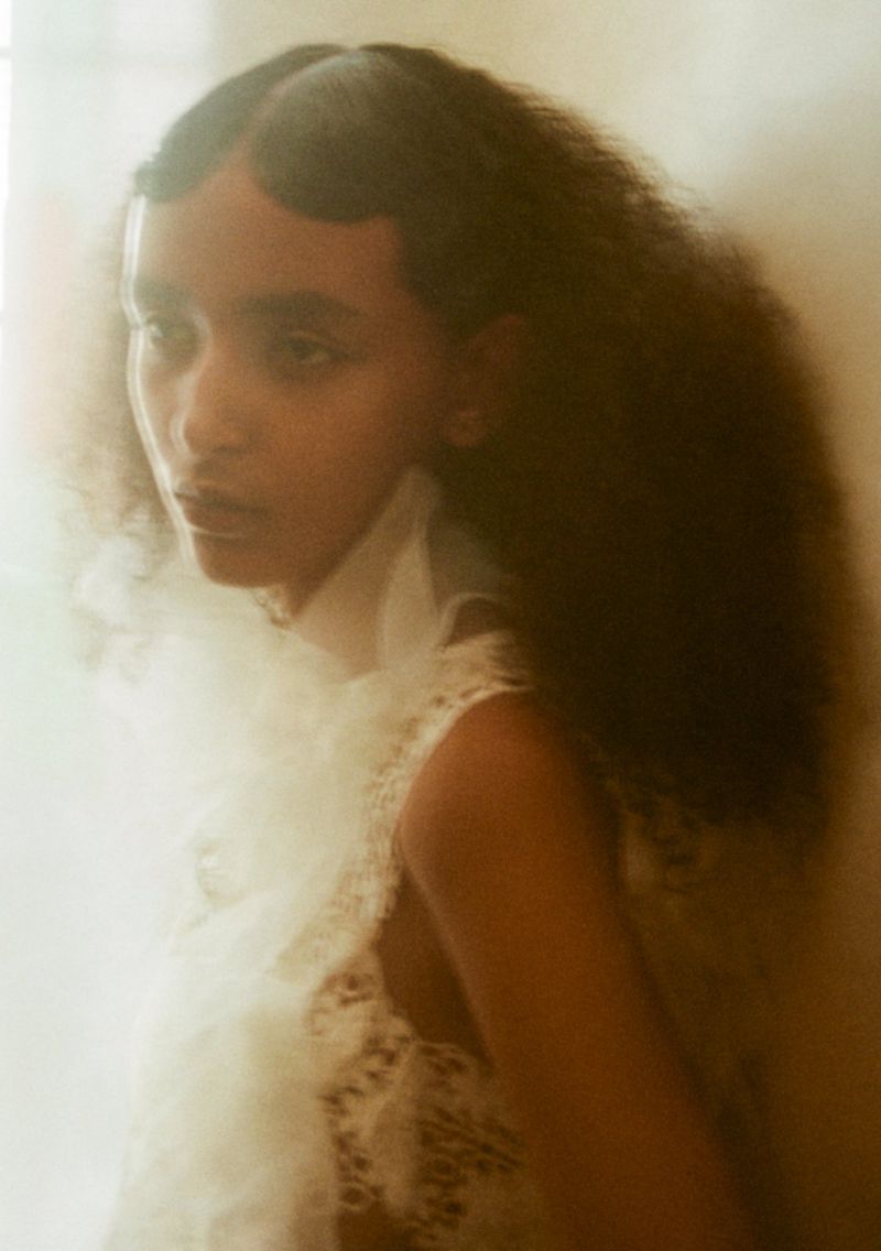 Portrait of an adult woman with dark curly hair wearing a white frilly garment, shot in soft, diffused lighting in an indoor setting.
