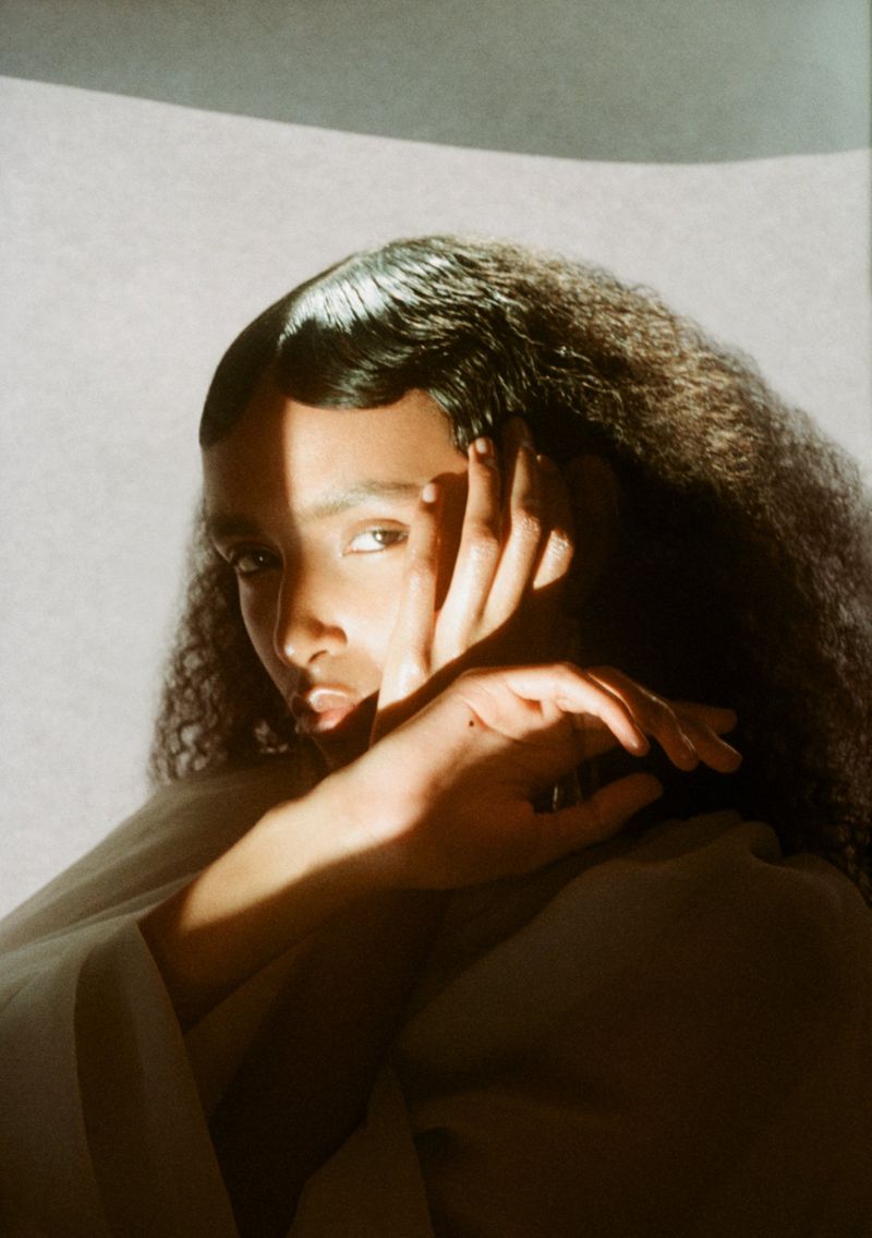 Portrait of a young woman with dark curly hair, partially lit by sunlight, with her hand near her face in a photo studio setting.