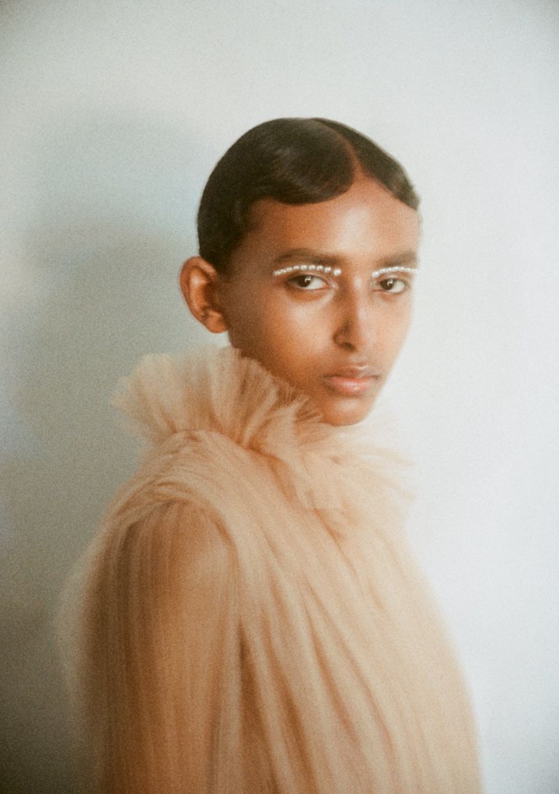 Close-up studio shot of a fashionable adult female with dark hair, a pale dress with a high frilled collar, and pearl eye makeup.
