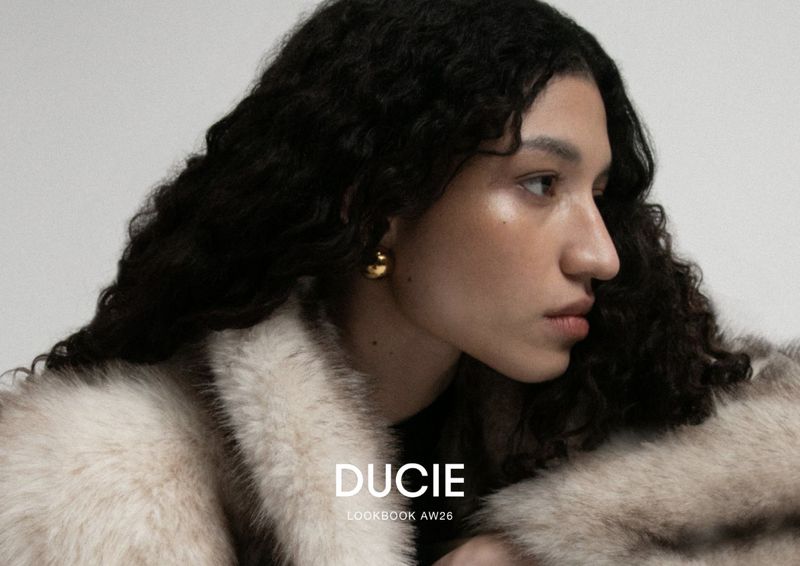 Model Keny Salgado wearing a beige fur coat and a spherical gold earring against a studio backdrop for Ducie.