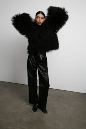 Model Keny Salgado wearing an oversized black shaggy jacket and wide-leg leather trousers against a white studio wall.