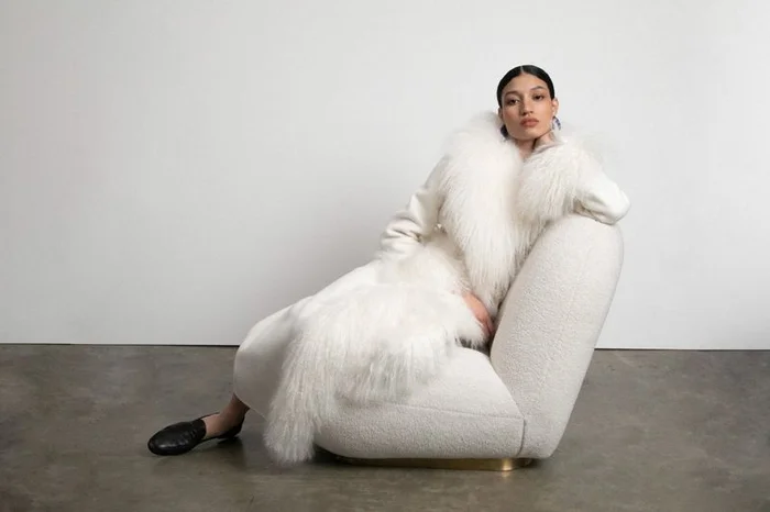 Model Keny Salgado wearing a plush white shearling coat while seated in a modern curved white armchair on a studio floor.