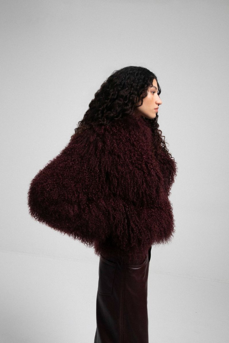 Model Keny Salgado posing in profile wearing a voluminous textured burgundy jacket and dark trousers against a grey studio backdrop.