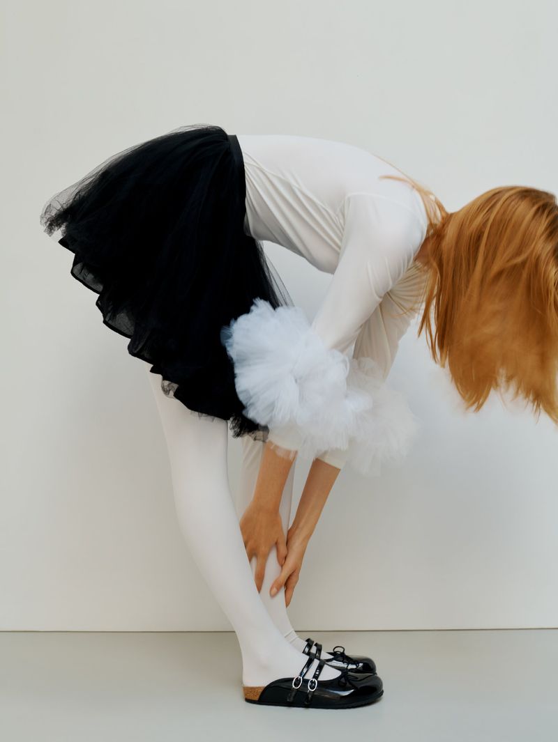 Woman in a black tulle tutu, white top, white tights, and black buckle shoes bending forward against a white studio wall.