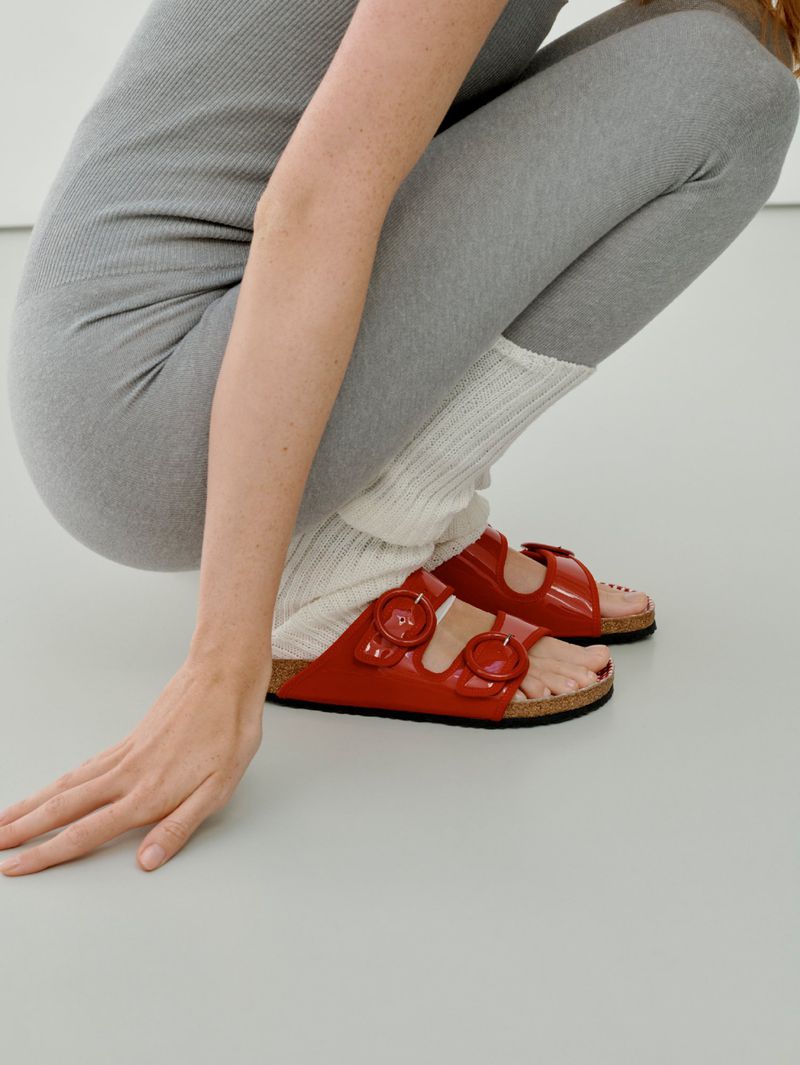 A person crouching down wearing glossy red Birkenstock sandals with cork footbeds and cream-colored ribbed leg warmers over grey leggings.