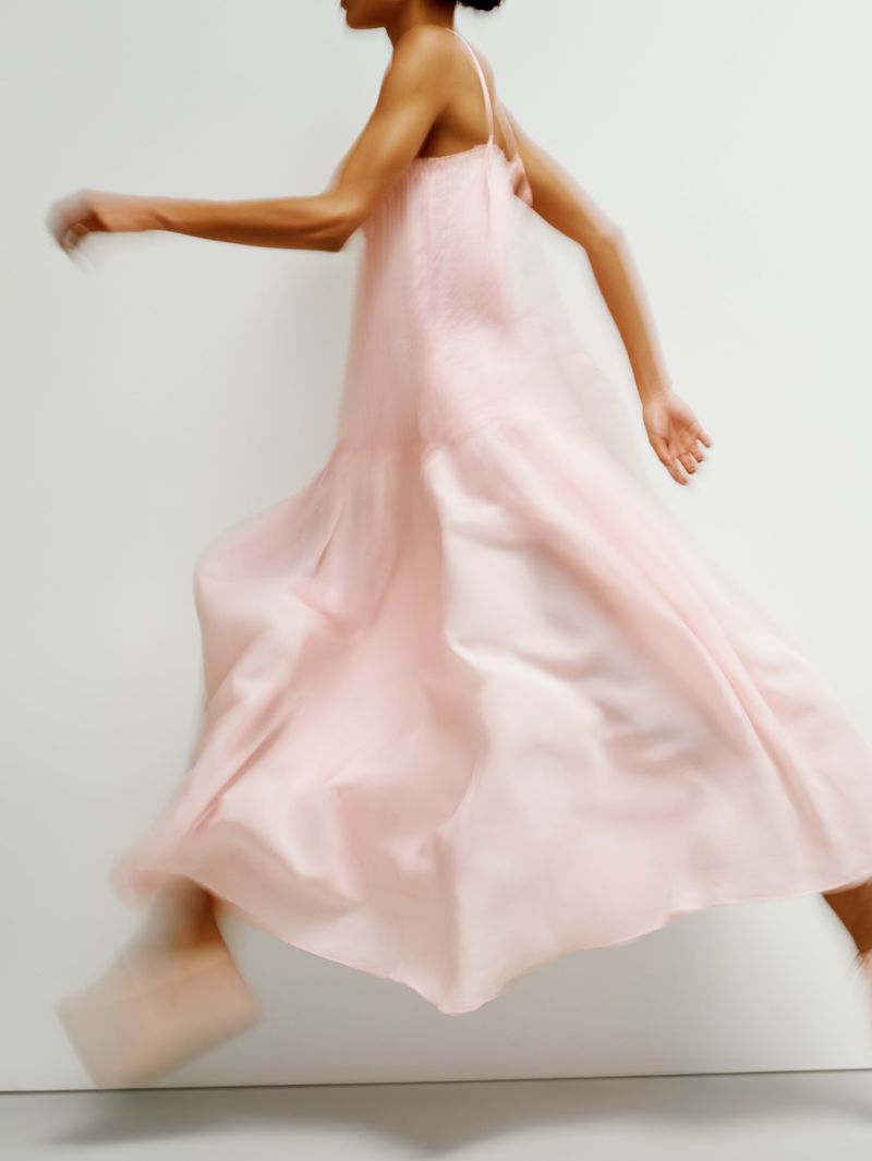 Motion-blurred model leaping across a white studio in a voluminous pastel pink spaghetti-strap dress.