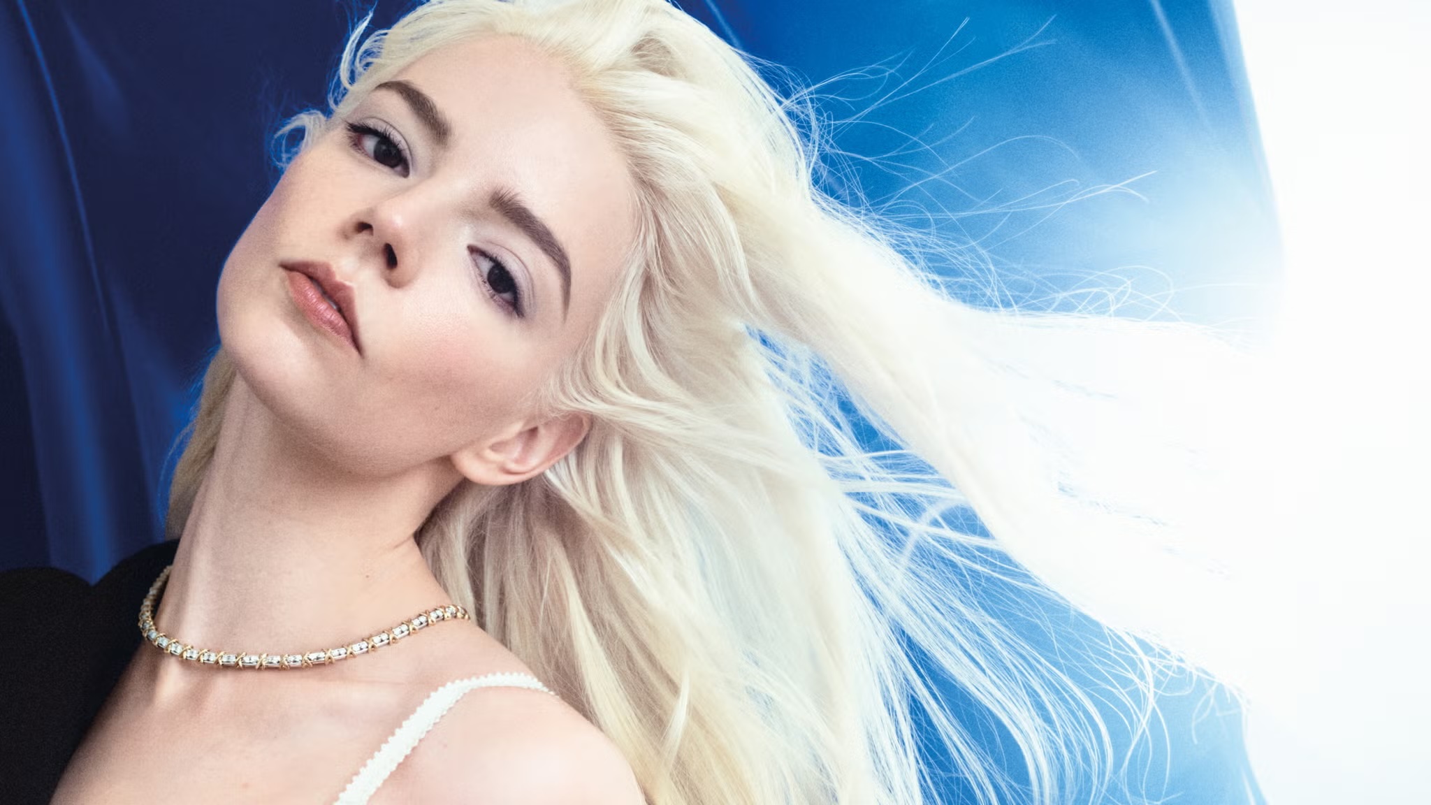 Portrait of Anya Taylor-Joy with platinum blonde hair and a necklace against a blue and white background