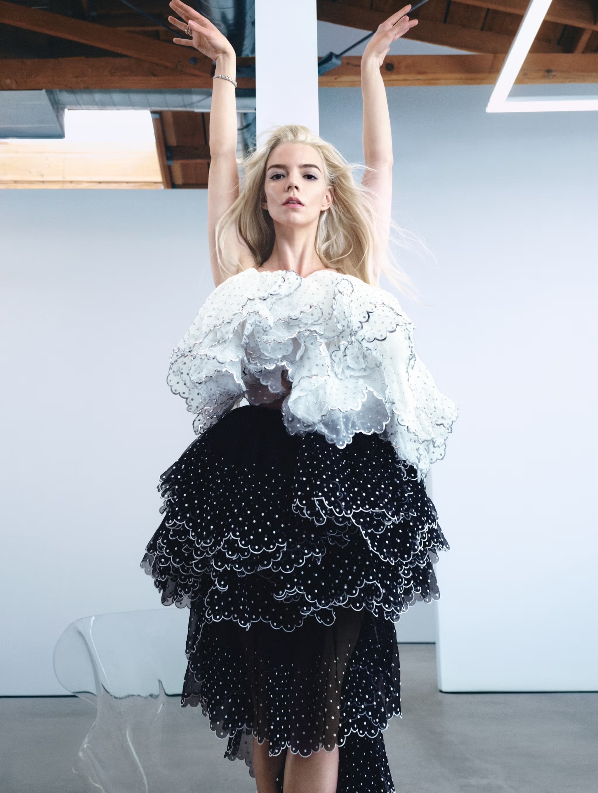 Anya Taylor-Joy posing with arms raised overhead wearing a dramatic black and white ruffled polka dot dress in a bright studio.
