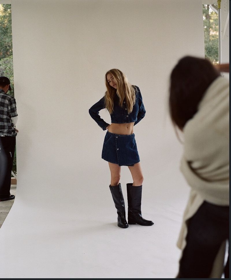 Model Thea Ros posing in a dark denim cropped jacket, mini skirt, and tall black boots on a white seamless backdrop on set.