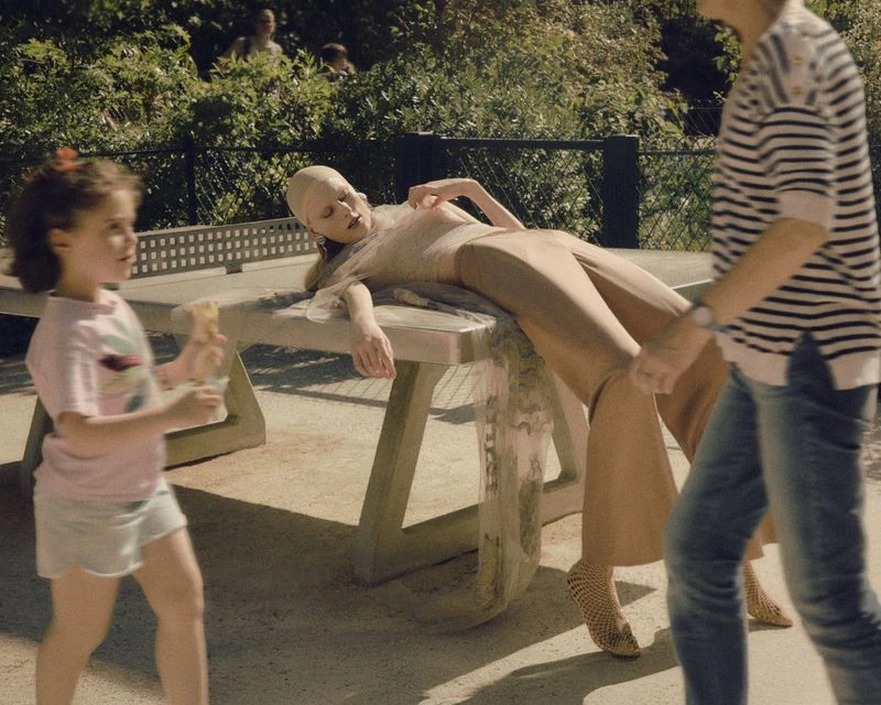 A bald adult lays on a concrete table outside while a toddler approaches and an adult walks by on a sunny day in a natural setting.