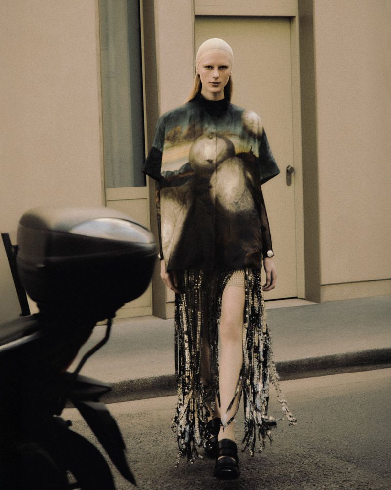 Fashion model in artistic tunic and fringe skirt walks on urban street beside parked scooter, showcasing unique designer style and dark color palette.