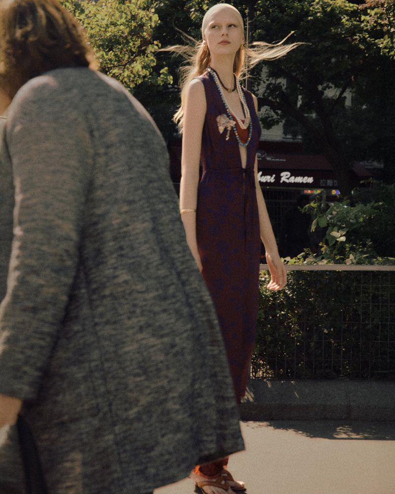 A woman in a burgundy dress walks on a city street near a restaurant, flanked by two blurred pedestrians under sunny, natural lighting.