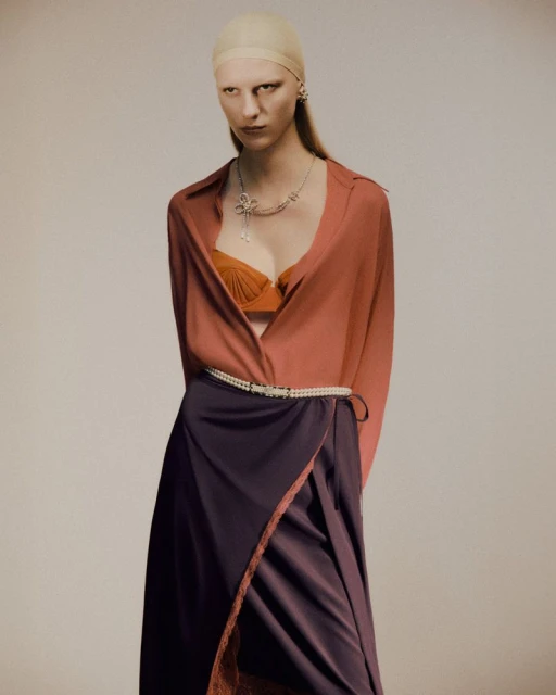 A woman with fair skin, a cap, and jewelry poses in a studio wearing an orange bra, an open rust-colored shirt, and a purple satin skirt.