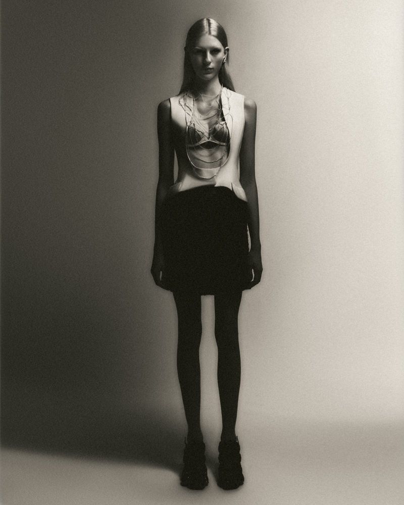 Monochrome fashion shot of a slender woman in a light sleeveless top, dark mini skirt, and chunky boots, standing against a gray backdrop.
