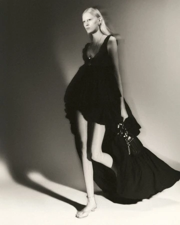 Monochrome fashion shot of a woman with pale complexion wearing a dark, tiered, sleeveless dress, and silver sandal-like shoes against a wall.