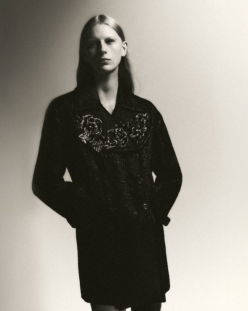 Model Eduarda Muehlmann wearing a textured black coat with floral embroidery on the collar in a monochrome studio portrait.