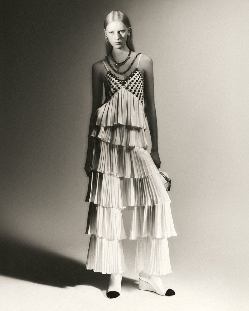 Monochrome fashion shot: fair-skinned woman in layered, tiered dress and matching boots, jewelry, holding purse, against gradient backdrop.