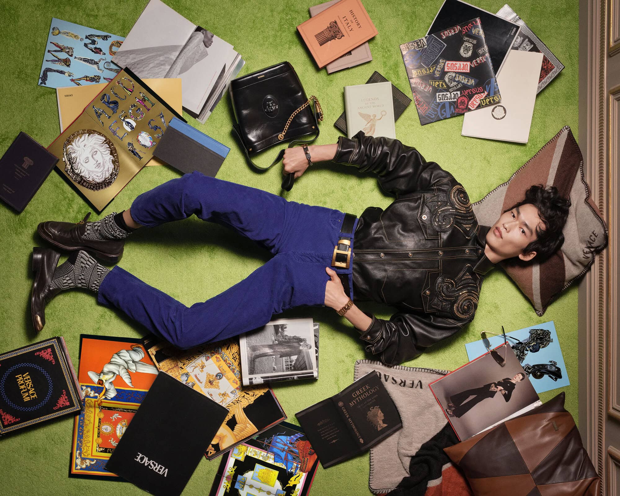East Asian model lying on a bright green surface in a black leather jacket and blue pants, surrounded by art books and pillows.