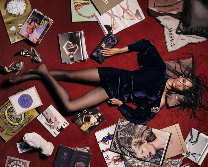 Model lying on a deep red carpet in a navy velvet mini dress surrounded by fashion magazines, high heels, and Versace accessories.