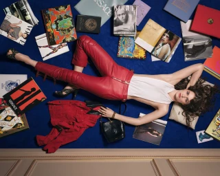 Model lies across a royal blue carpet wearing red leather trousers and a white halter top, surrounded by open fashion books and magazines.