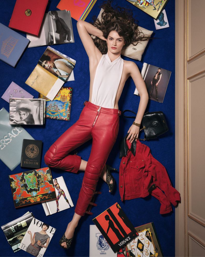 Model lying on a deep blue carpet surrounded by books and fashion magazines, wearing a white halter top and red leather trousers.