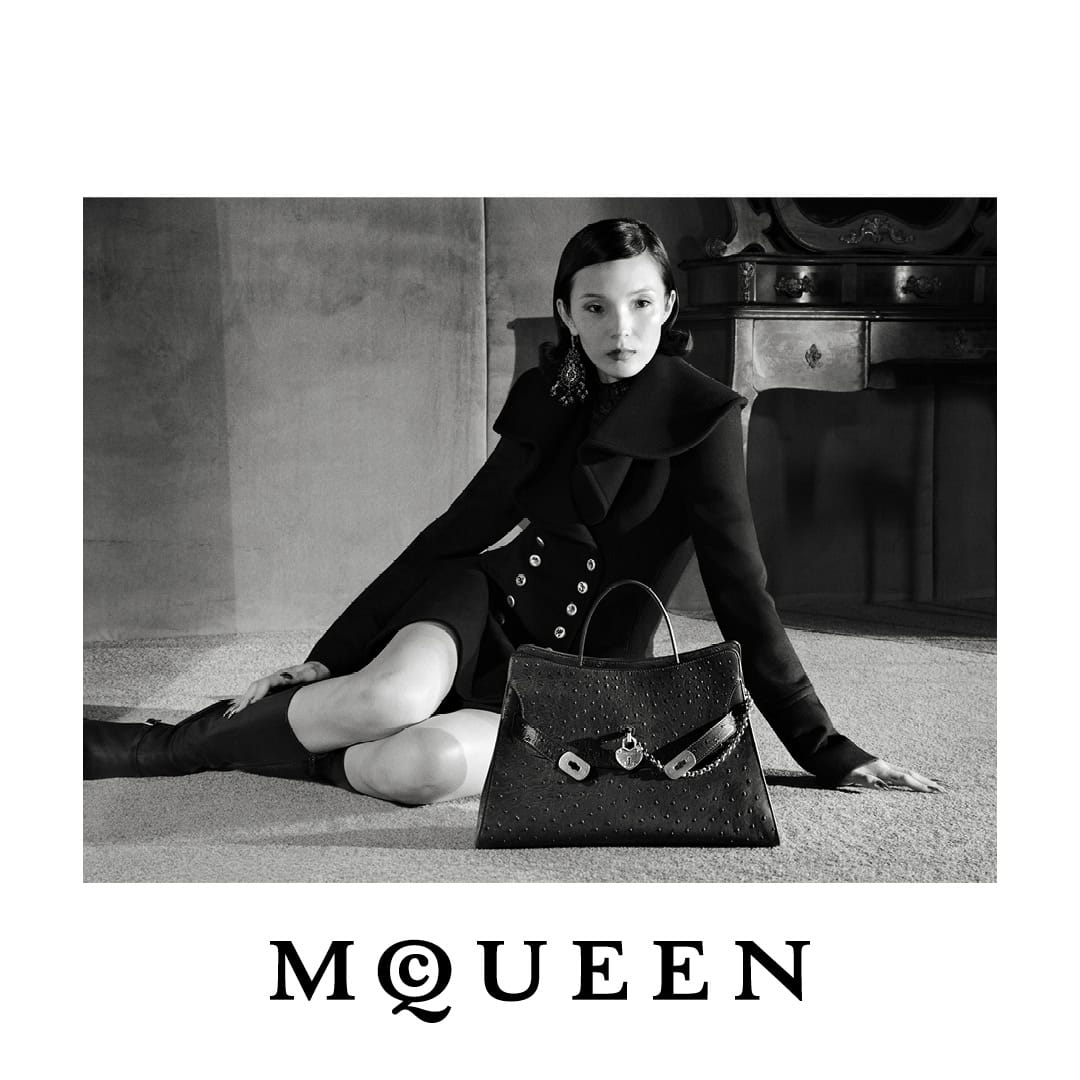 Model Xiao Wen Ju wearing a black tailored coat seated on the floor beside a black leather McQueen handbag in black and white.