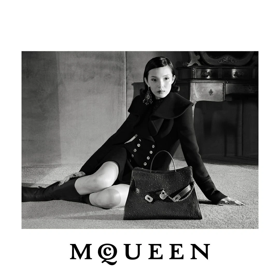 Model Xiao Wen Ju wearing a black tailored coat seated on the floor beside a black leather McQueen handbag in black and white.