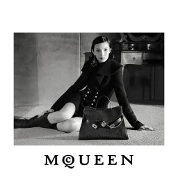 Model Xiao Wen Ju wearing a black tailored coat seated on the floor beside a black leather McQueen handbag in black and white.