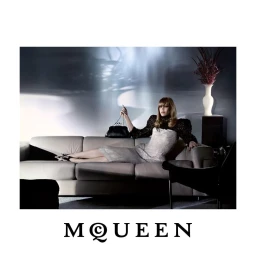 Karen Elson reclines on a beige leather sofa holding a black clutch bag in the McQueen Fall 2026 advertising campaign.