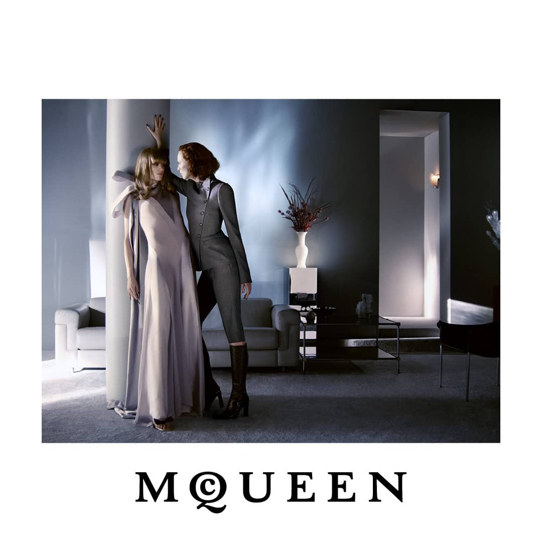 Two models posing by a column in a moody interior, one in a draped pale lavender gown and the other in a tailored dark grey suit.