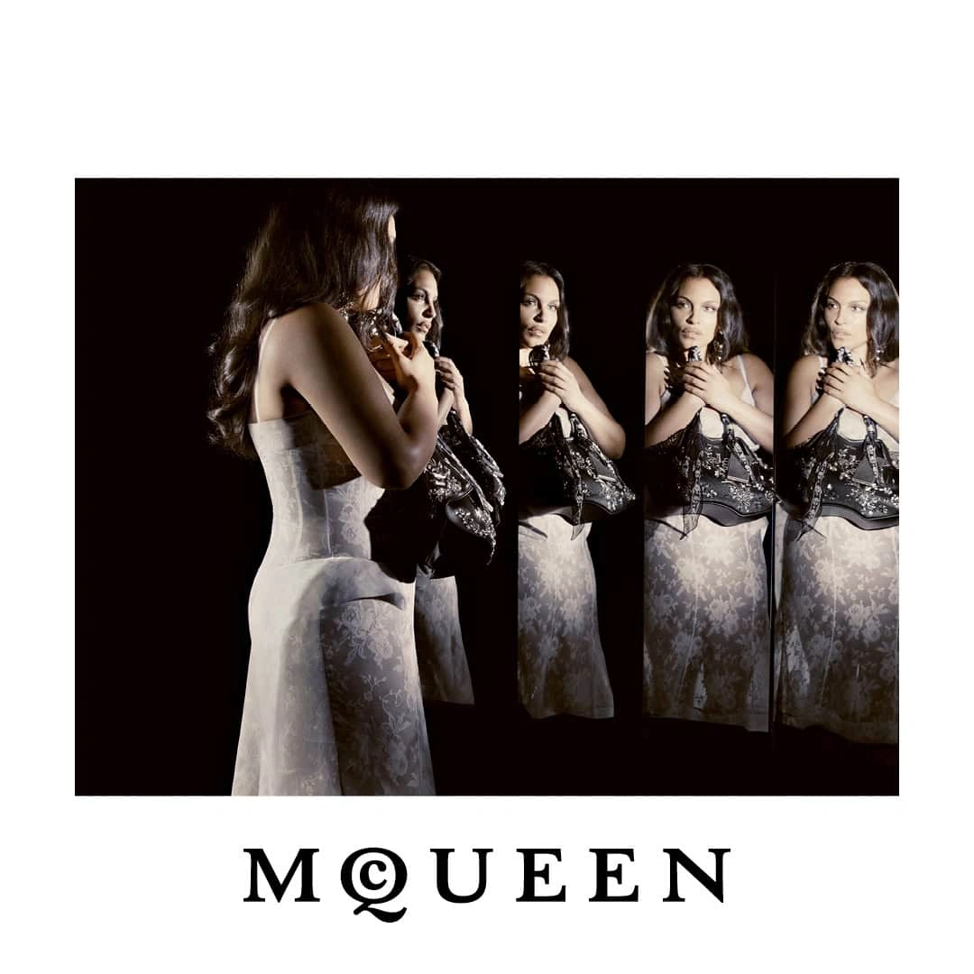 Paloma Elsesser in a white lace dress reflected in mirrors against a black backdrop for McQueen Fall 2026.
