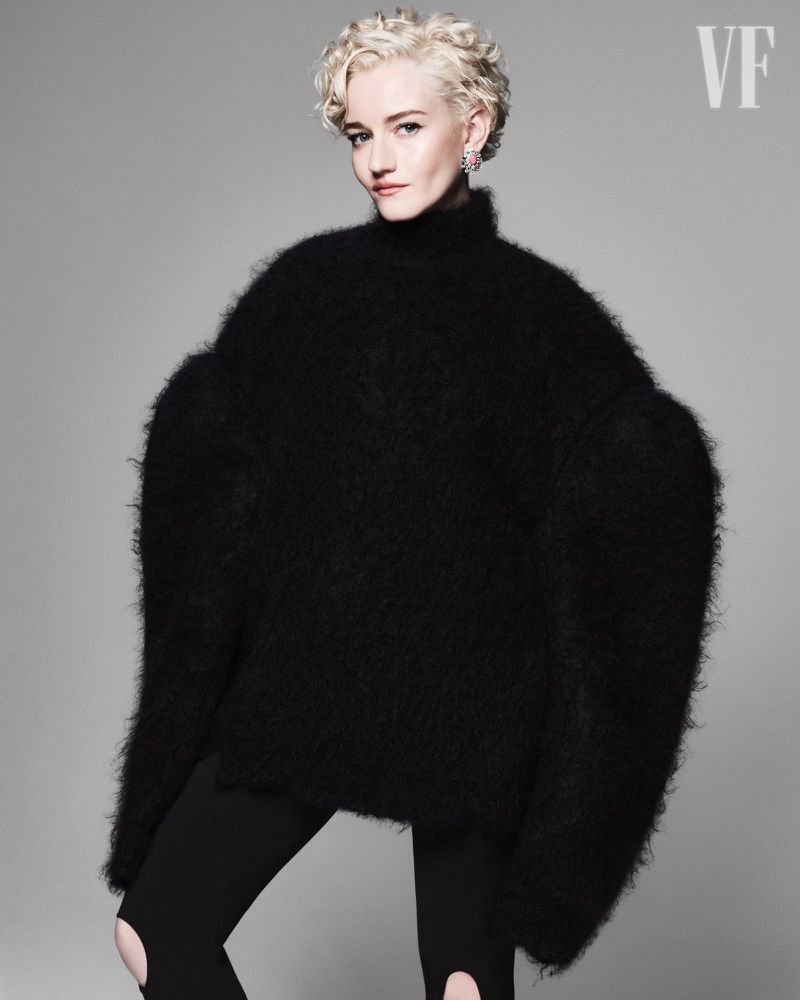 A woman with short blonde hair poses in a photo studio wearing a large black sweater and leggings with cutouts, against a gray background.