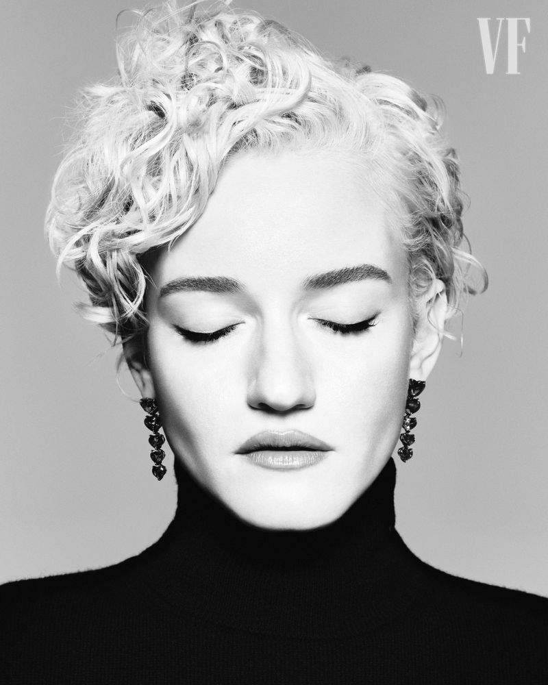 Black and white portrait of a woman with closed eyes, blonde curly hair, turtleneck, and dangling earrings against a gray backdrop.