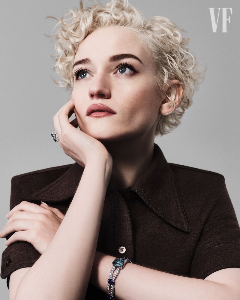 Elegant studio portrait of a young adult woman with wavy blonde hair wearing a brown collared shirt, jewelry, and looking upward.