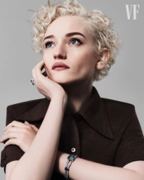 Portrait of actress Julia Garner with short blonde curly hair wearing a dark brown collared top against a grey seamless background.