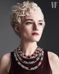 Julia Garner wearing layered ruby and diamond collar necklaces and a burgundy sleeveless top against a grey studio backdrop.