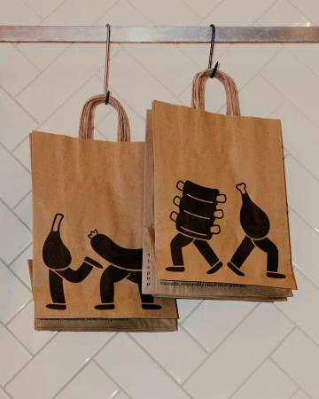 Two brown kraft paper bags with illustrated meat characters hanging from metal hooks against a white herringbone tiled wall.