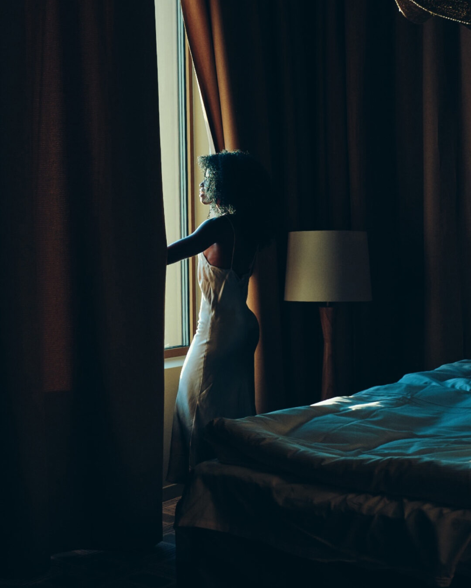 Woman in a white slip dress looking out a window framed by heavy dark curtains inside a dim hotel room.