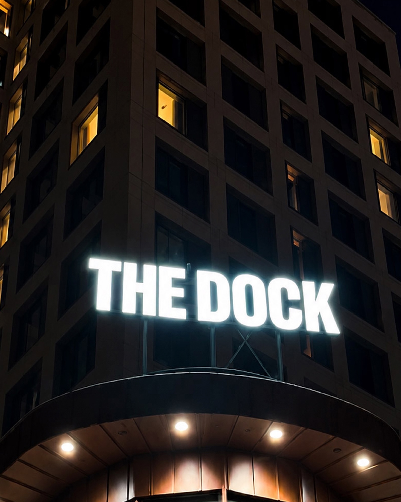 Glowing white neon sign reading THE DOCK mounted on a dark building facade with illuminated windows at night.