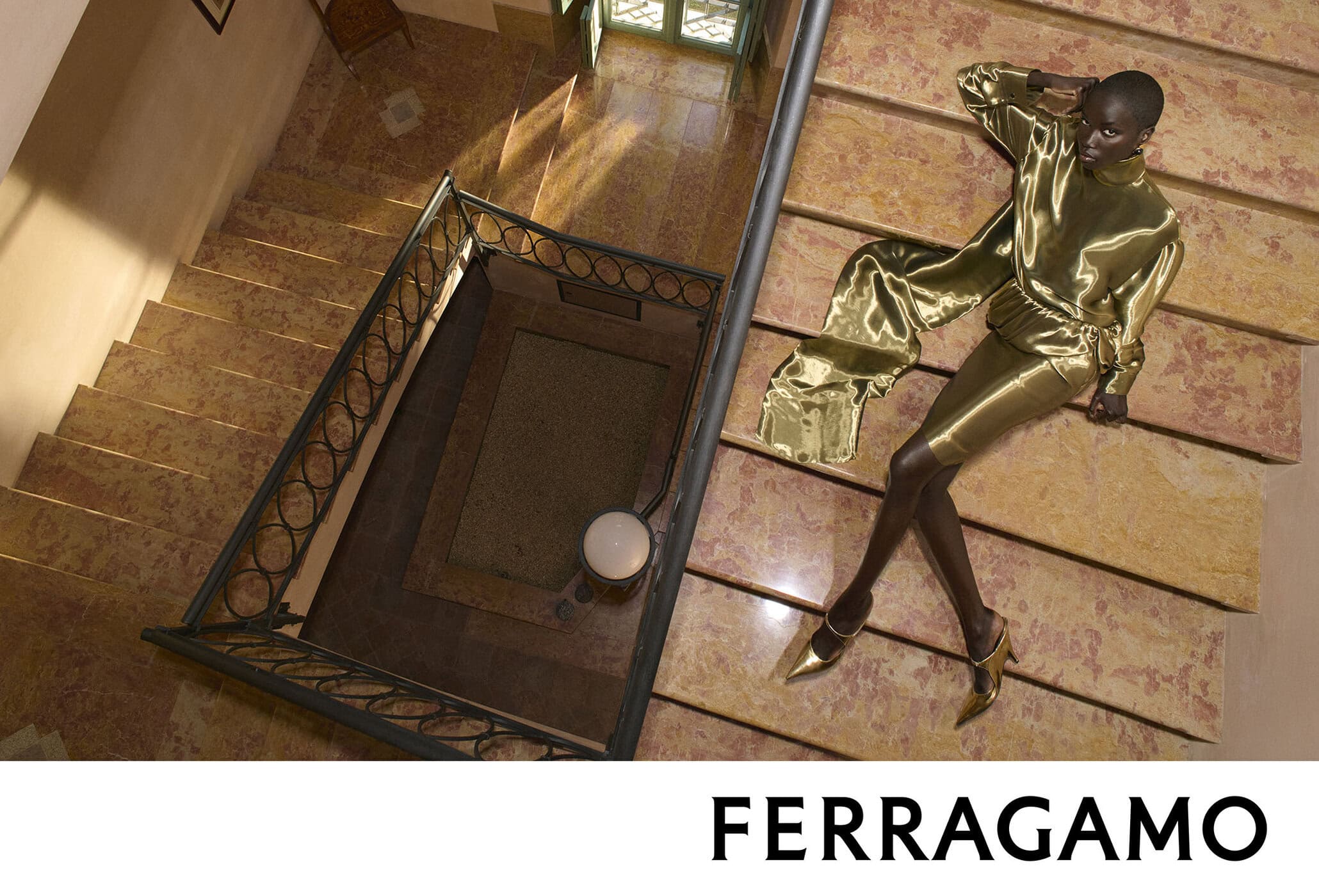Model wearing a glossy metallic gold outfit reclining on a grand marble staircase for Ferragamo Fall 2026 ad campaign.