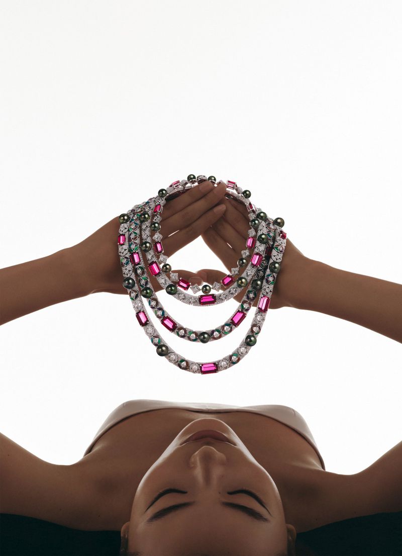 Adult woman lying on her back, holding up layered pearl and gemstone necklaces; jewelry commercial, lit against a bright white background.