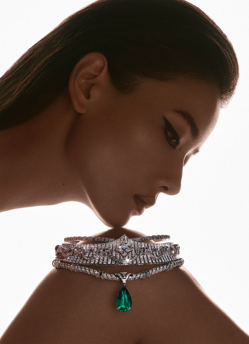 Profile view of a woman wearing a diamond and emerald choker and tiara against a white background in a fashion-style beauty shot.