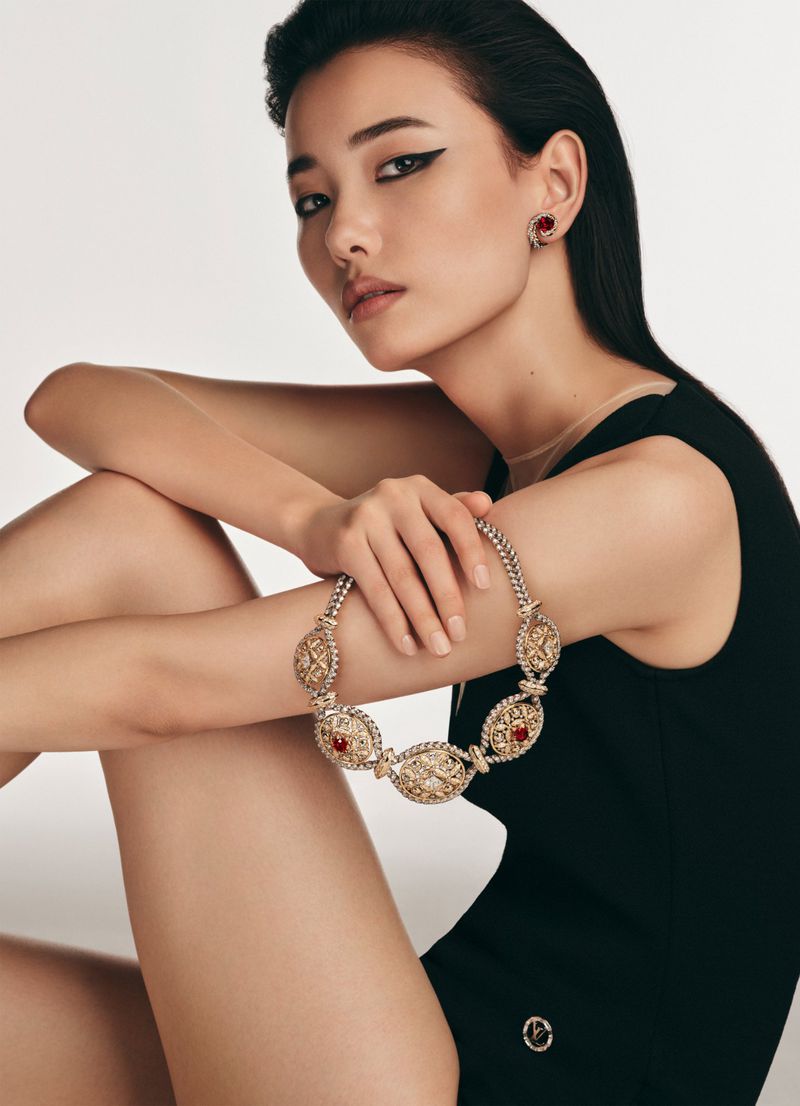 Elegant Asian woman poses indoors wearing a black dress and red jewelery necklace, highlighting studio-style fashion and beauty.