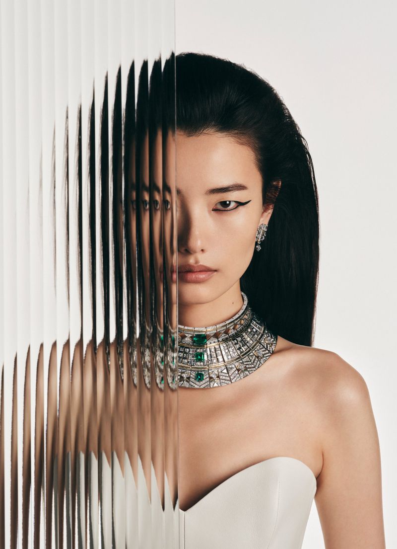 Asian woman with black hair and ornate necklace poses in a studio portrait with a textured glass panel in the foreground.