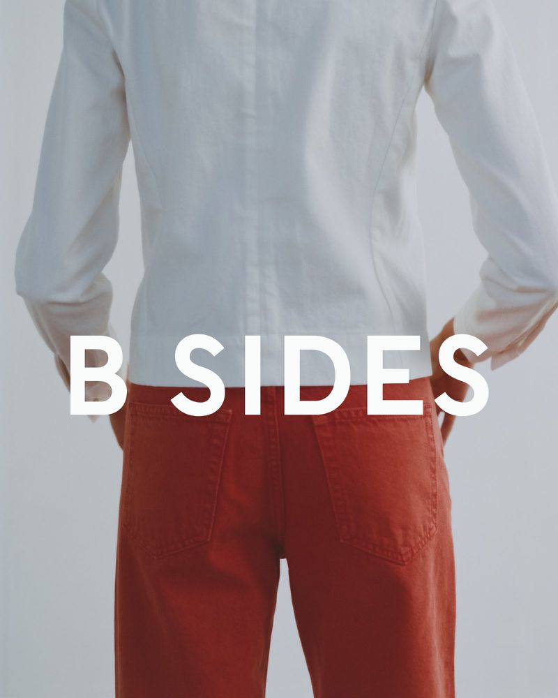 Model seen from behind wearing a white long-sleeve shirt and rust orange denim pants with white B SIDES text overlay.