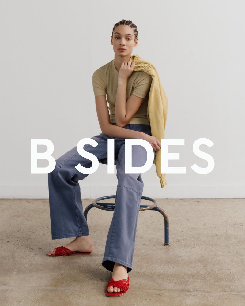 Model Aliza Jarmon seated on a stool wearing a beige t-shirt, wide-leg blue jeans, and red slide sandals.