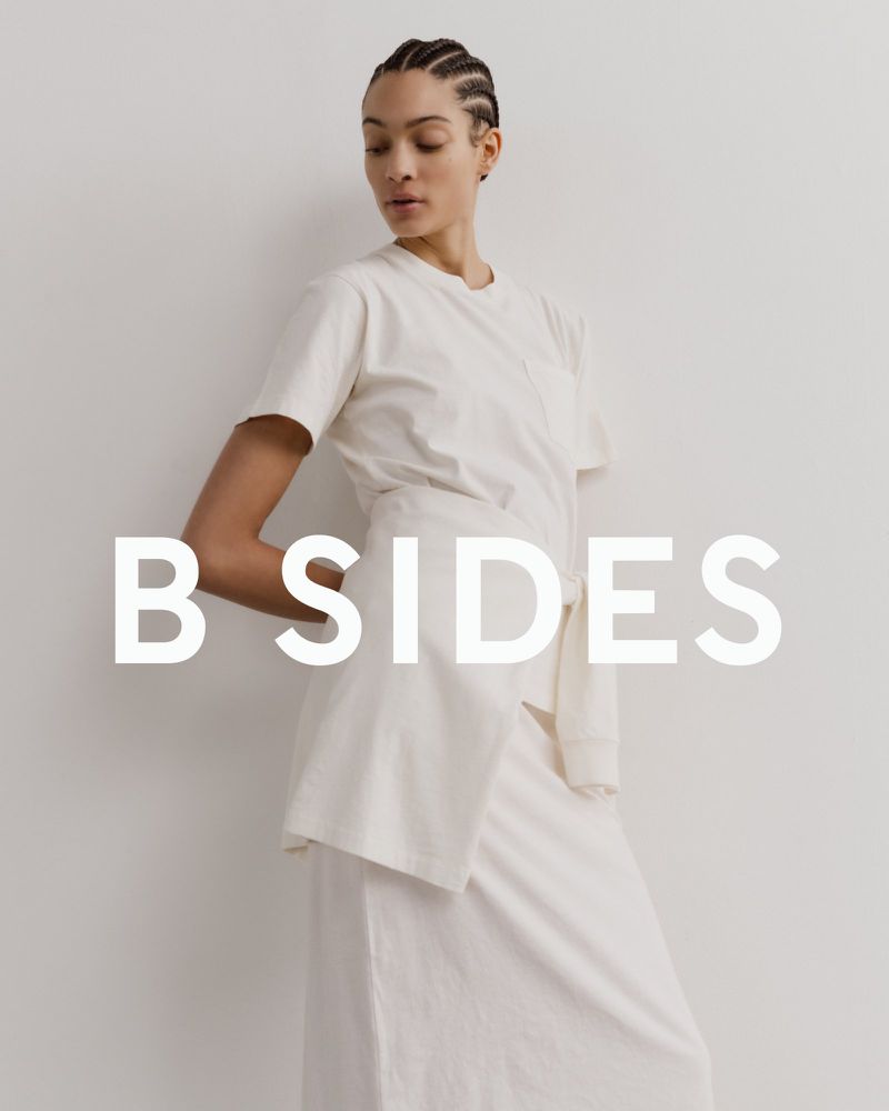 Model Aliza Jarmon wearing an off-white t-shirt and tied wrap skirt against a neutral backdrop with B SIDES text overlay.