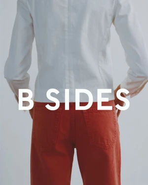 Model seen from behind wearing a white long-sleeve shirt and rust orange denim pants with white B SIDES text overlay.