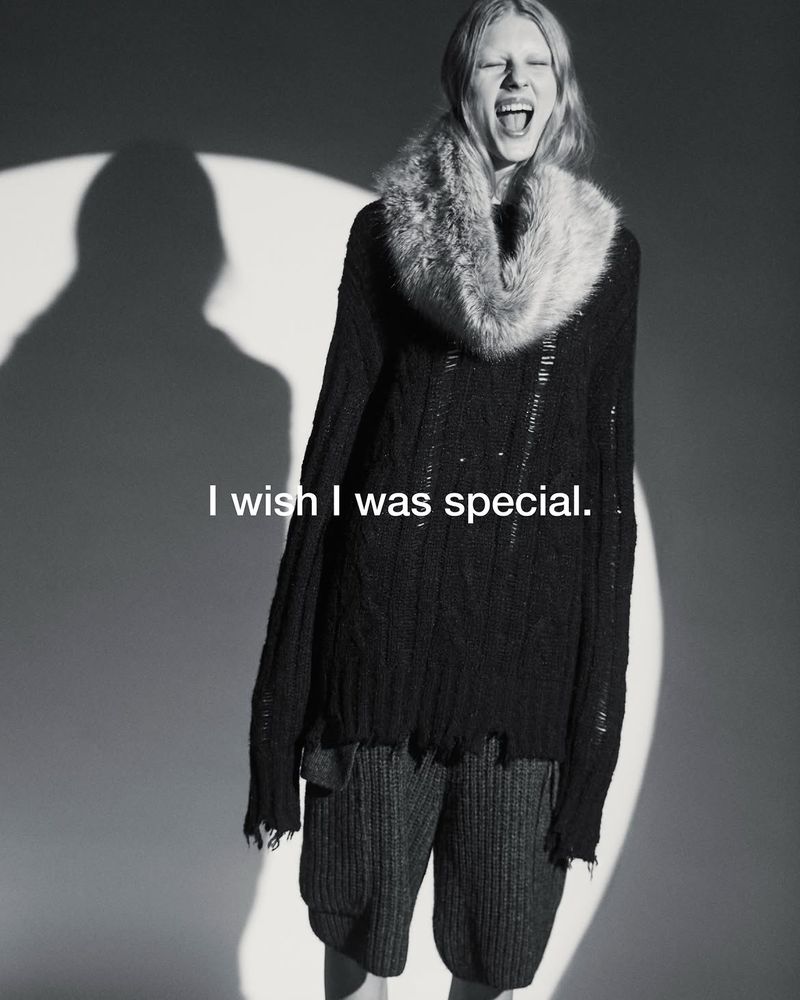 Model wearing an oversized distressed black knit sweater with a fur collar laughing against a circular spotlight backdrop.